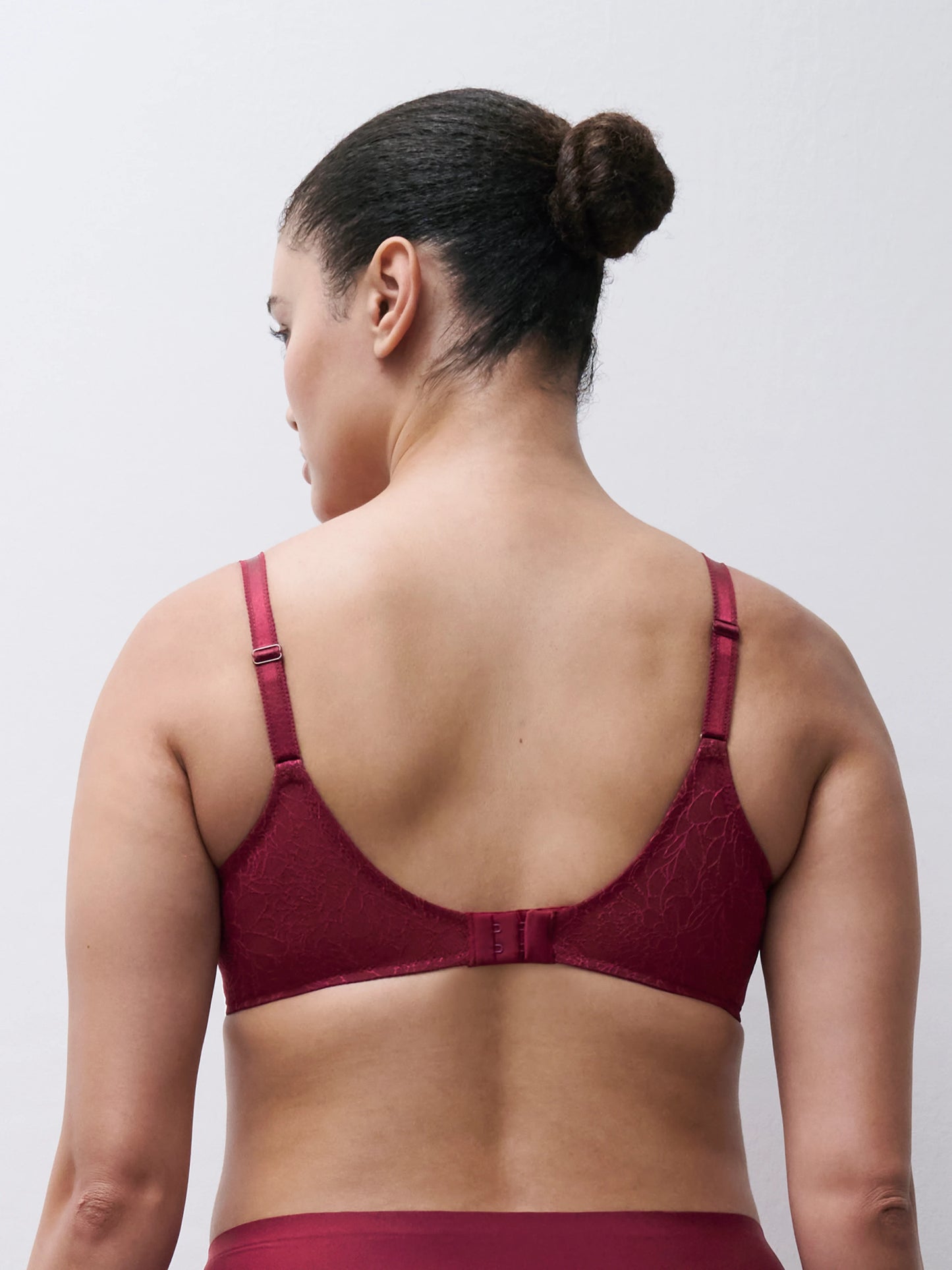 Rear view of a woman wearing a burgundy lace underwire bra against a light gray studio background.