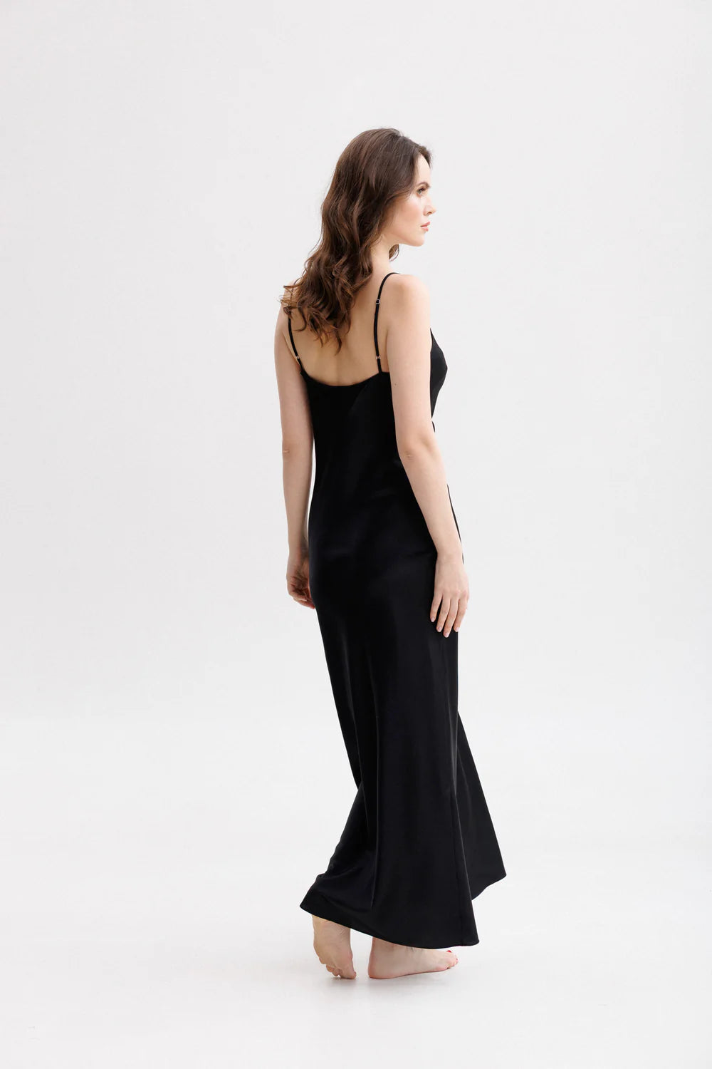 Woman wearing a long silk black slip dress on a spaghetti straps showing the back pose. 